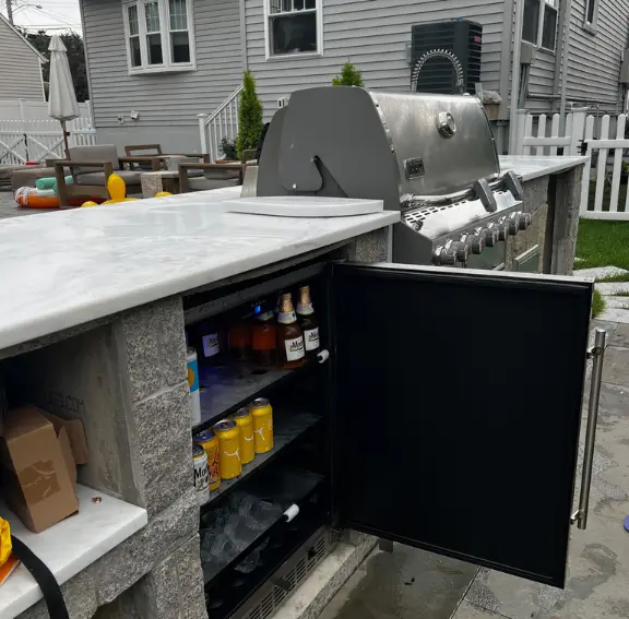 A modern outdoor kitchen design featuring an L-shaped island with a built-in stainless steel grill, beverage fridge, and weather-resistant cabinetry.